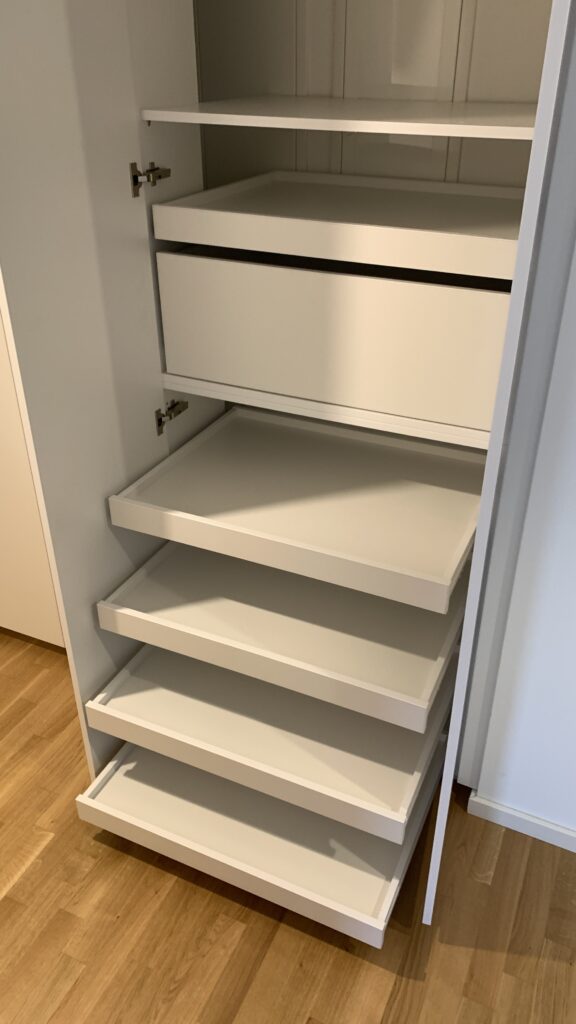 Ein offener weißer Kleiderschrank mit mehreren herausziehbaren Ablageböden und einem integrierten Schubfach. Die Böden sind leer, und der Schrank steht auf einem hellen Holzboden. Die Tür ist geöffnet, sodass die Innenstruktur gut sichtbar ist.