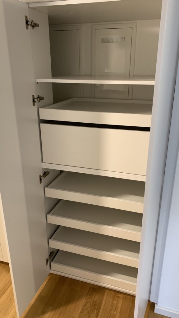 Ein offener weißer Kleiderschrank mit mehreren herausziehbaren Ablageböden und einem integrierten Schubfach. Die Böden sind leer, und der Schrank steht auf einem hellen Holzboden. Die Tür ist geöffnet, sodass die Innenstruktur gut sichtbar ist.